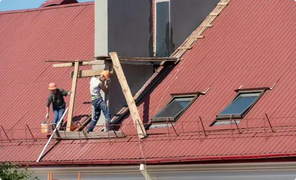 Two roofers on wooden scaffolding installing metal accents on a red metal roof — workers compensation coverage
