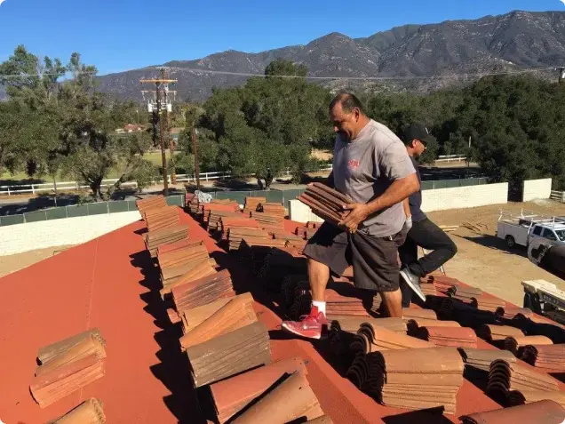 Two roofers carrying and laying bundles of red clay tile on a sloped roof with mountains in the background